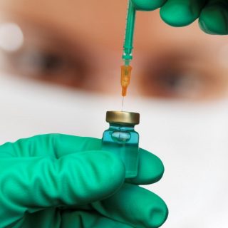 Hand in Gummischuhen zieht mit Spritze Impfstoff aus einem Fläschchen