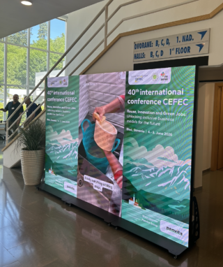 Große digitale Anzeigetafel im Foyer eines Konferenzgebäudes mit dem Plakat zur ‚40th international conference CEFEC – Reuse, Innovation and Green Jobs: Unlocking inclusive business models for the future‘ in Bled, Slowenien; im Hintergrund sind ein Treppenaufgang, Wegweiser und einzelne Personen zu sehen.
